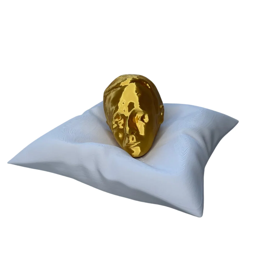 Escultura de un rostro dorado apoyado sobre un cojín blanco nacarado, impresa en 3D